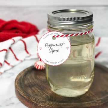 A glass mason jar full of off-white, clean peppermint syrup with a red and white striped thread wrapped around the mason jar neck with a circular white tag that says, "Peppermint Syrup".
