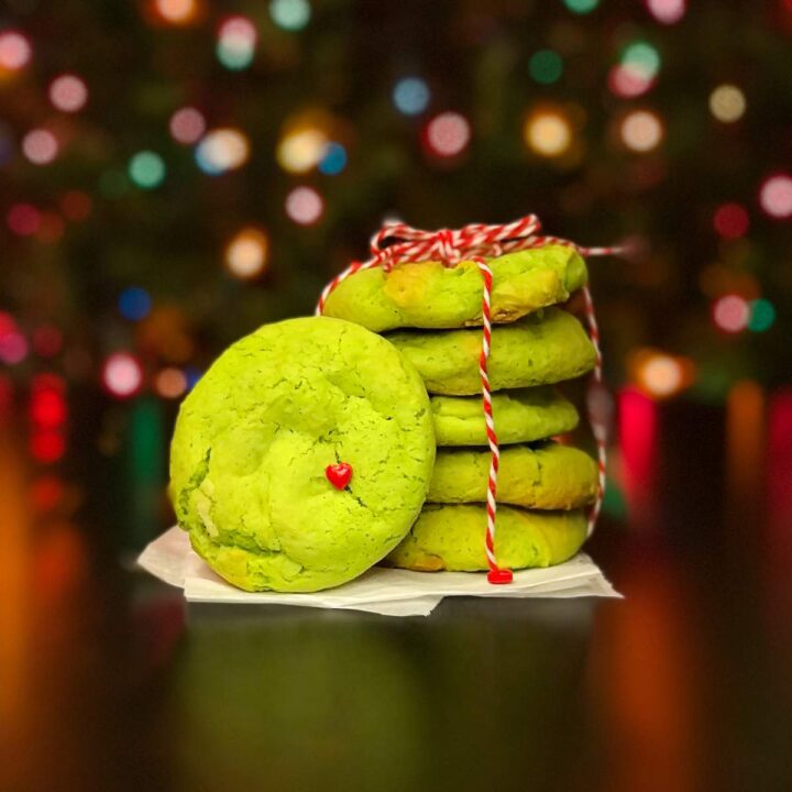 A bright green cake mix cookie with a small red heart attach leans against a stack of neon green cookies tied together with red and white twine in front of a Christmas tree with rainbow Christmas lights.