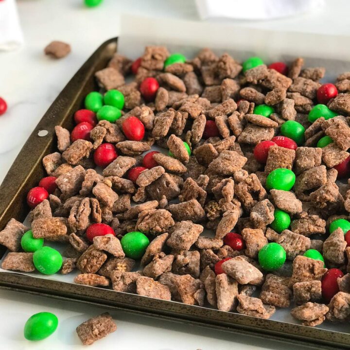 Christmas Muddy Buddies with light brown, chocolate coated cereal squares and red and green round peanut holiday m&m candies on a parchment sheet that is lined with a sheet of white parchment paper.