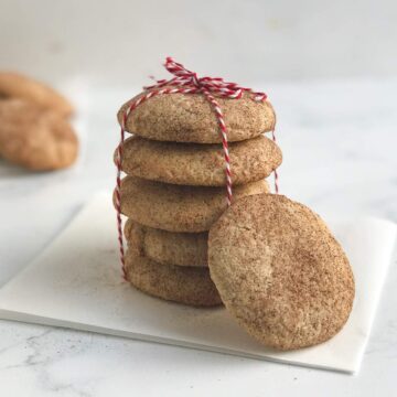 A round, cinnamon sugar coated snickerdoodle cake mix cookie leans on a stack of cake mix snickerdoodle cookies tied in red and white twine.