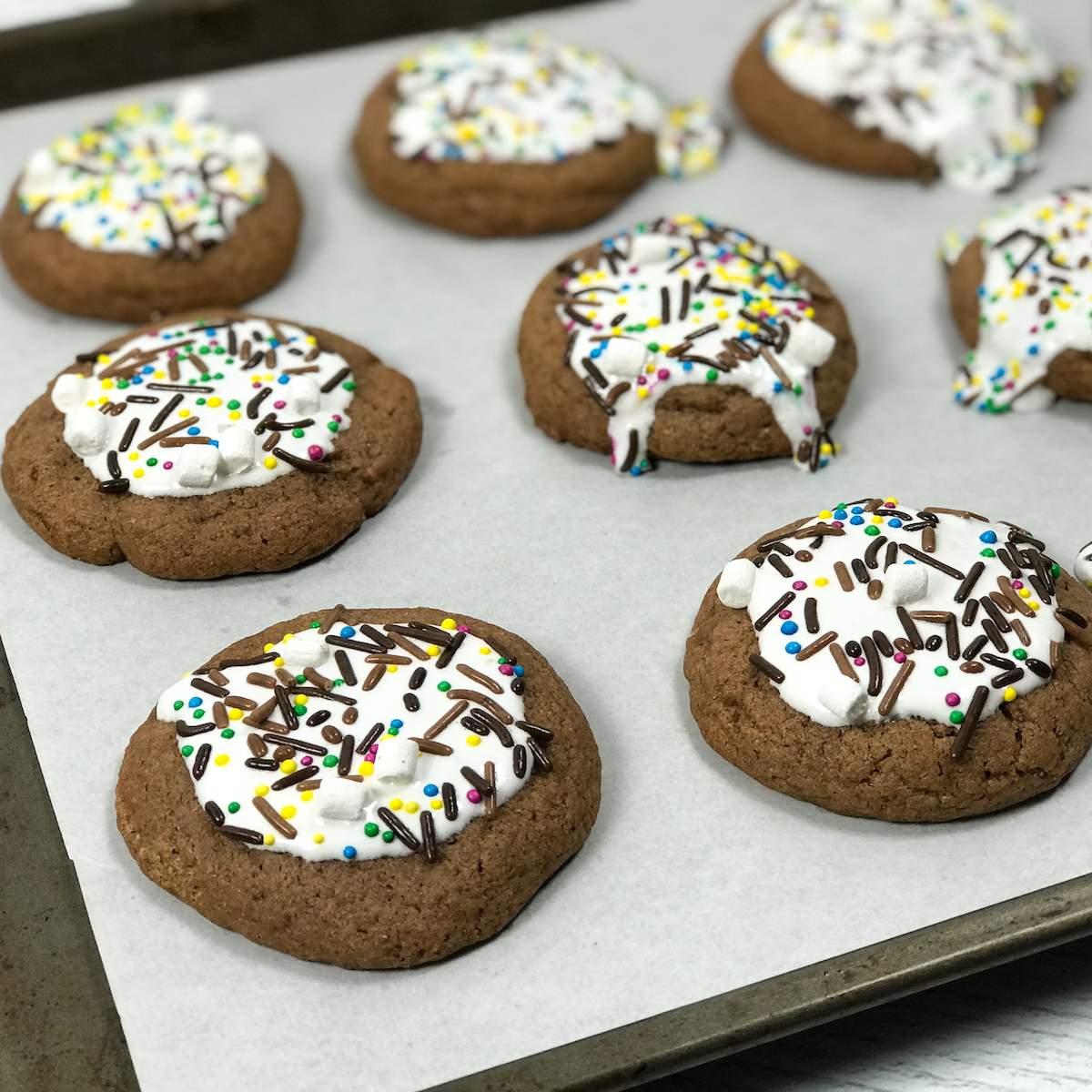 Dark brown hot cocoa sugar cookies with bright white marshmallow creme icing and hot chocolate sprinkles lay on a baking sheet that is lined with parchment paper.