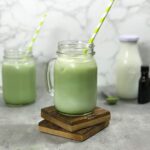 A beautiful green vanilla matcha latte in a glass mason jar with a handle sits on top wooden coasters in front of another beautiful green matcha drink and the three ingredients in this Vanilla Matcha Latte recipe.