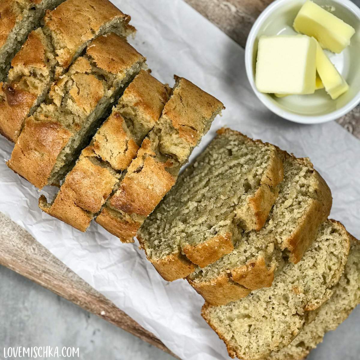 A sliced loaf of golden brown, soft banana bread with baking powder on a white sheet of parchment paper on a light brown wooden cutting board. 