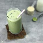 A glass of creamy, bright green matcha protein shake sits next to the three ingredients in this matcha protein shake recipe - a scoop of beige vanilla protein powder, a silver teaspoon of bright green matcha powder, and a milk bottle full of white milk.
