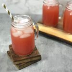 Beautiful, dark pink guava white tea lemonade sits in a mason jar glass with a handle on top of two dark brown wooden coasters. Two more beautiful dark pink iced guava tea lemonades, in mason jar glasses, sit in the background.