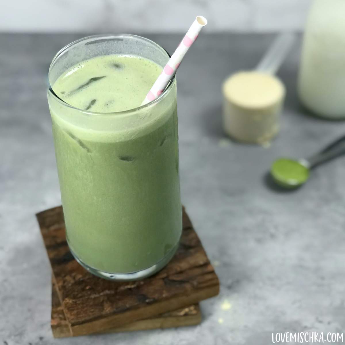Creamy, green matcha protein shake sits in a tall glass with ice cubes and a white and light pink polka dot paper straw.