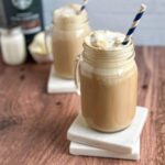 A light brown coffee milkshake with a cream foamy top in a mason jar glass with a handle with a blue and white striped paper straw.