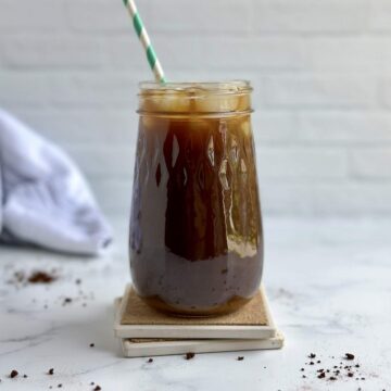 A dark brown iced coffee in a mason jar glass with a white and mint green stripped straw sits on two wooden coasters, surrounded by black instant coffee granules.