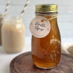 A mason jar bottle full of golden brown vanilla coffee syrup with a circular, light pink label that reads, "Vanilla Syrup".