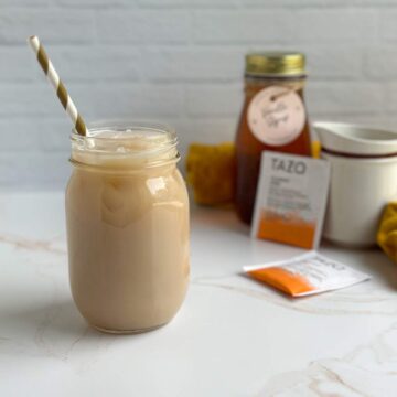 A tan, iced vanilla chai latte in a mason jar with a gold and white striped paper straw sits next to two chai tea bags, a mason bottle of homemade vanilla syrup for coffee, and a small container of creamy milk.