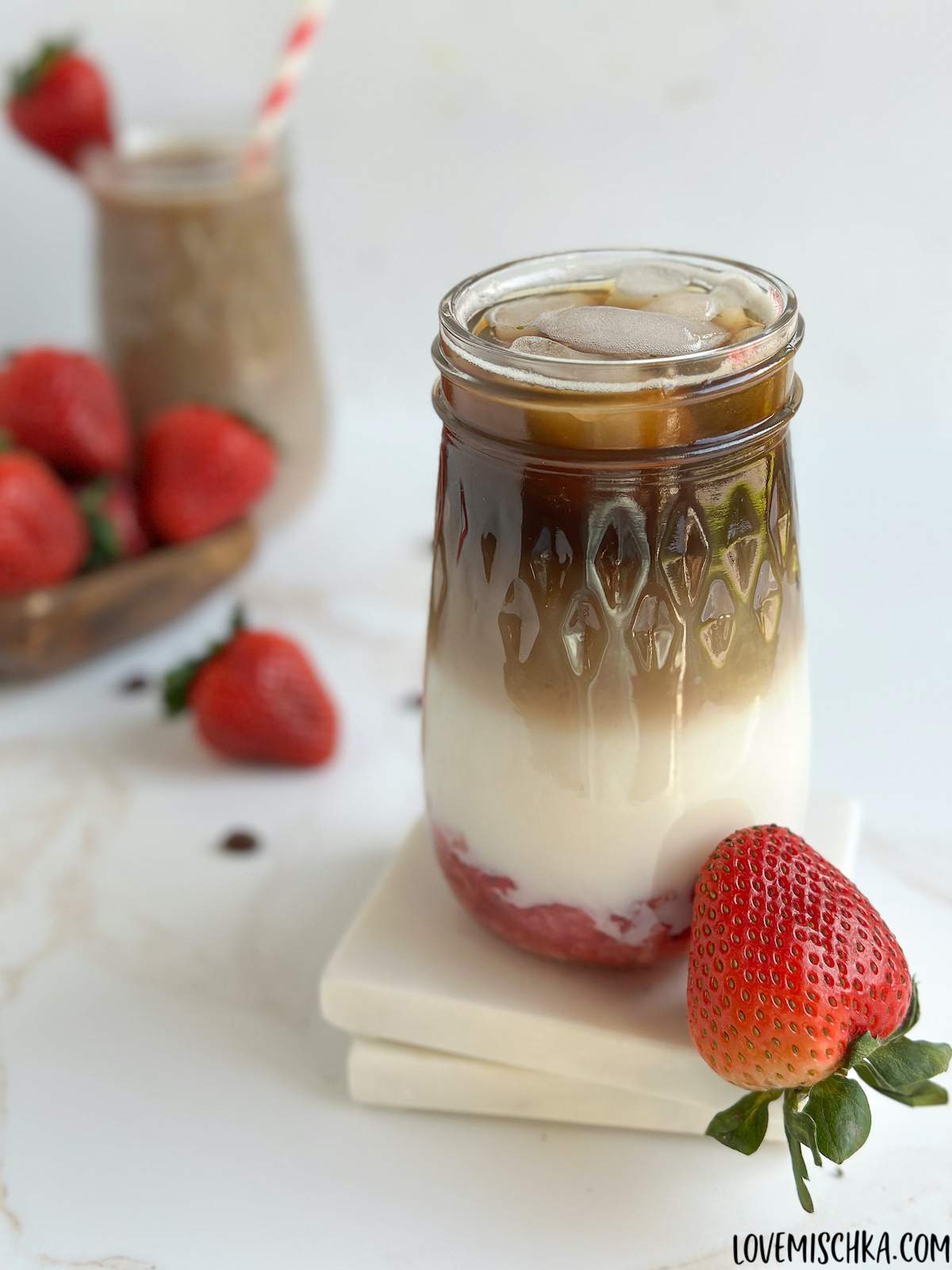 A layered strawberry latte in a tall mason jar glass with a bright red, juicy strawberry sitting against it with more bright red strawberries and another strawberry latte in the back. 