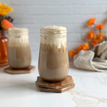 A brown, iced chai latte in a decorative mason jar with velvety pumpkin cold foam on top sits in between a beige dish towel with bright orange flowers and another chai drink topped with fluffy pumpkin cold foam.