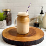 A layered iced oat milk latte in a mason jar glass with a handle and a purple and white striped paper straw.