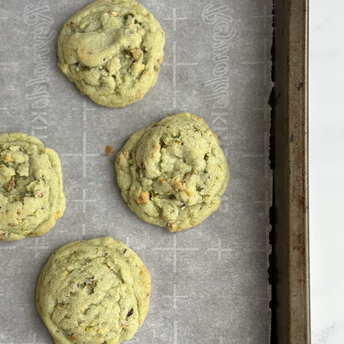 Three soft, chewy, bright green pistachio cookies lay on a sheet of white parchment paper on a used cookie sheet.