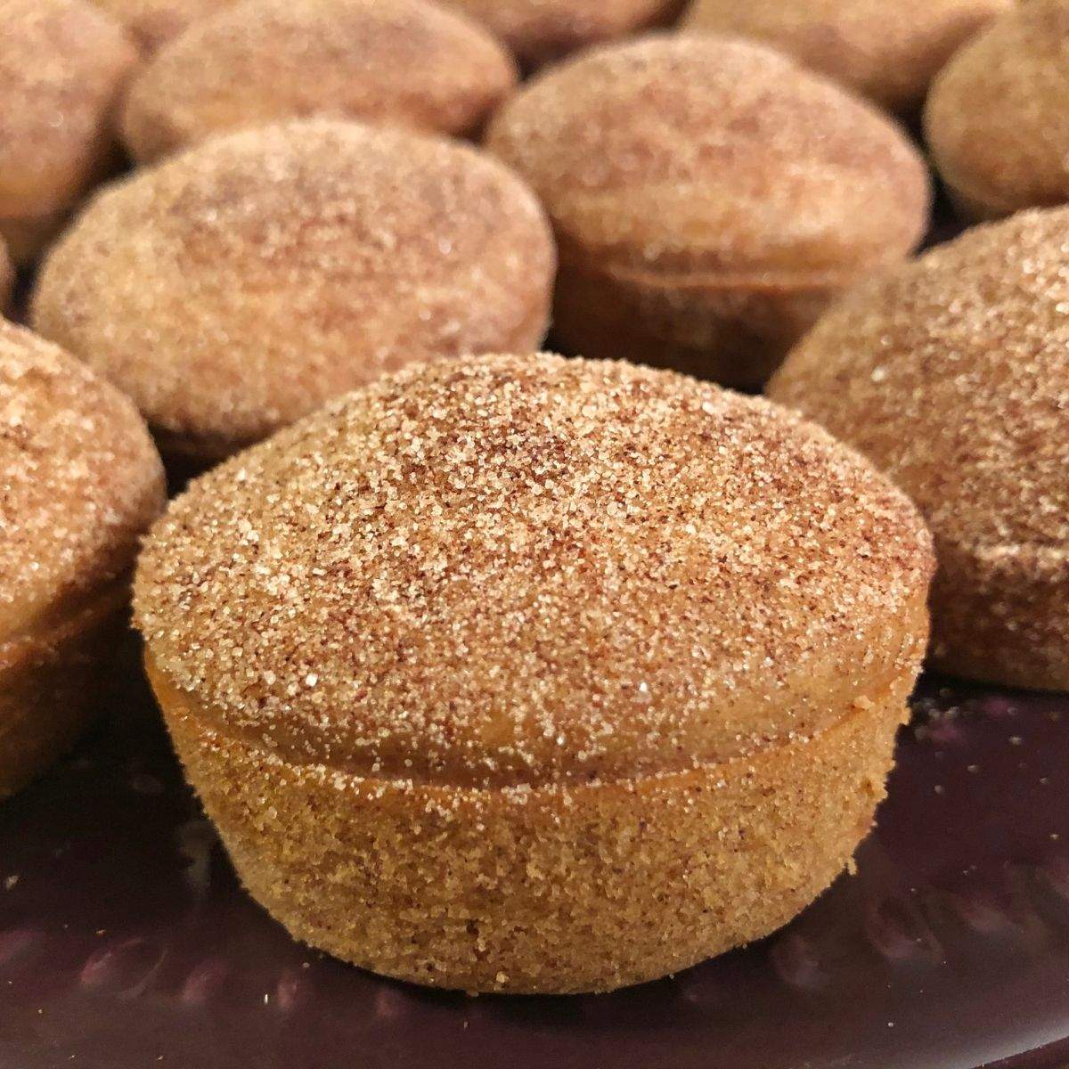 A close up of one Apple Cider Muffin, coated in cinnamon sugar, on a purple plate with lots of similar brown, cinnamon sugar coated muffins behind him.