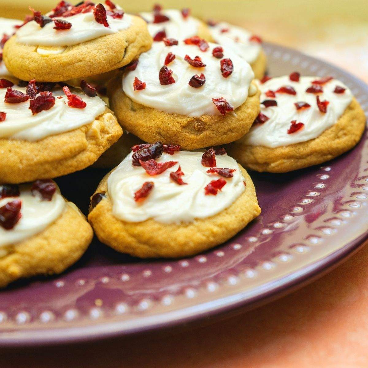 Beige Cranberry Bliss Bar Cookies with white, creamy frosting and small chunks of dried cranberries sit on a purple serving plate.