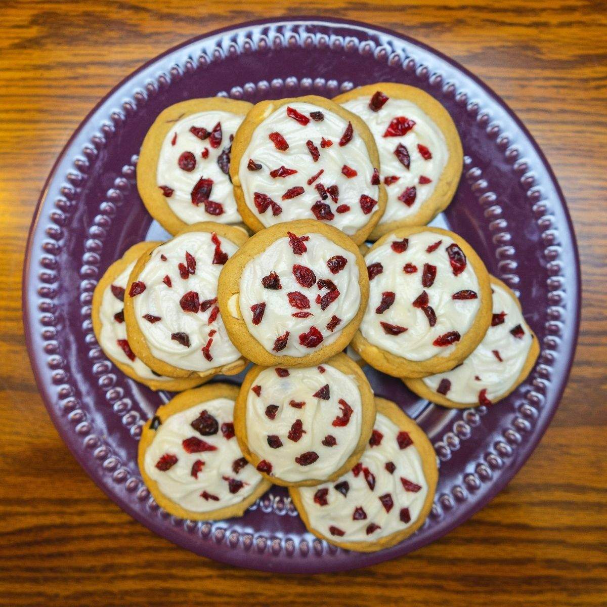 A purple plate of round beige cranberry bliss bar cookies that are topped with creamy, cream cheese-like frosting and small pieces of red dried cranberries.