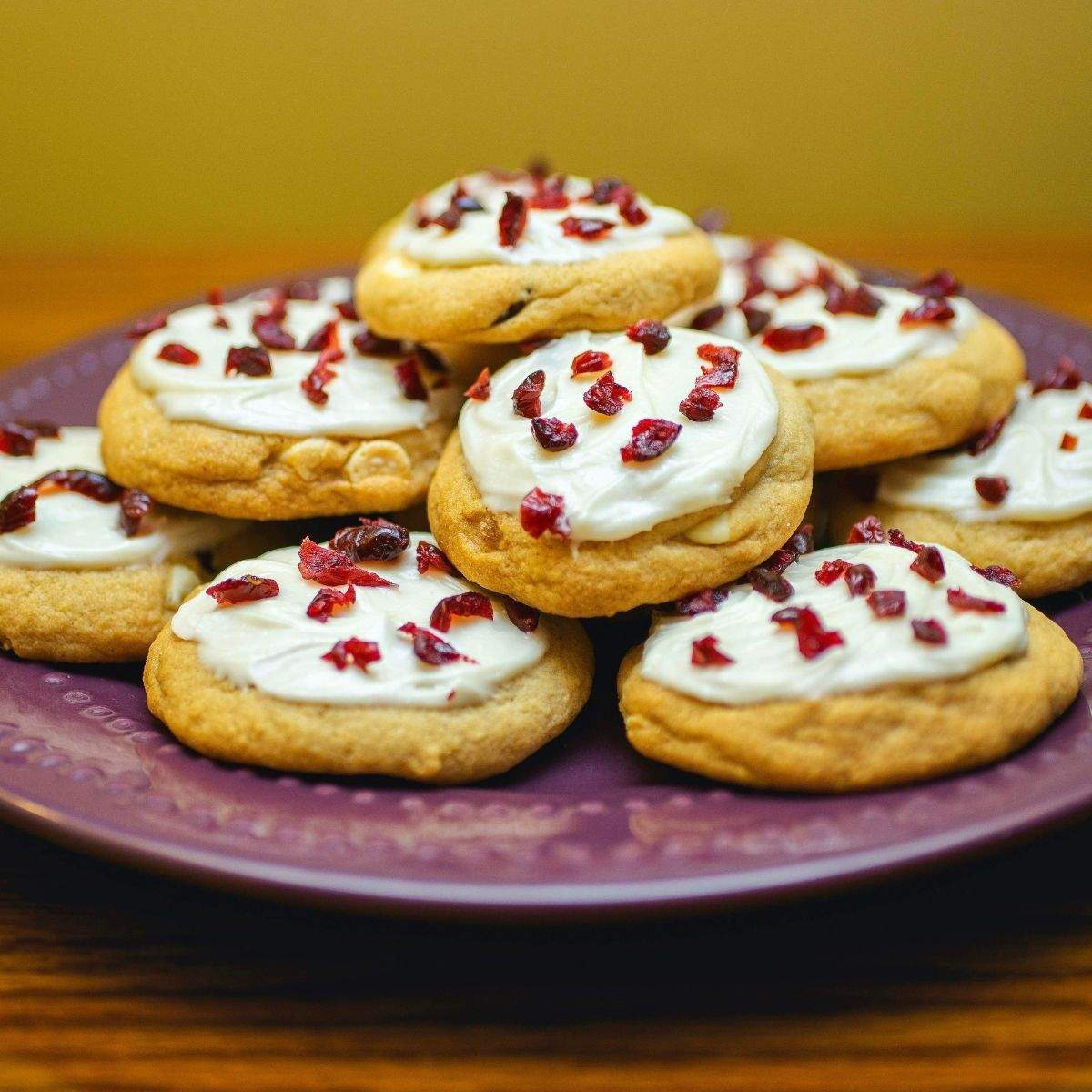 A stack of round tan Cranberry Bliss Cookies, topped with creamy, white frosting and pieces of dark red dried cranberries, on a round, purple serving plate.