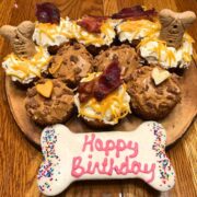 Ten pupcakes (cupcakes for dogs) sit on a circular wooden serving grey. Some cupcakes are topped with peanut butter and chewy dog treats. Others are topped with white cream cheese and shredded cheese. Crispy bacon and tan milkbones decorate the tops.