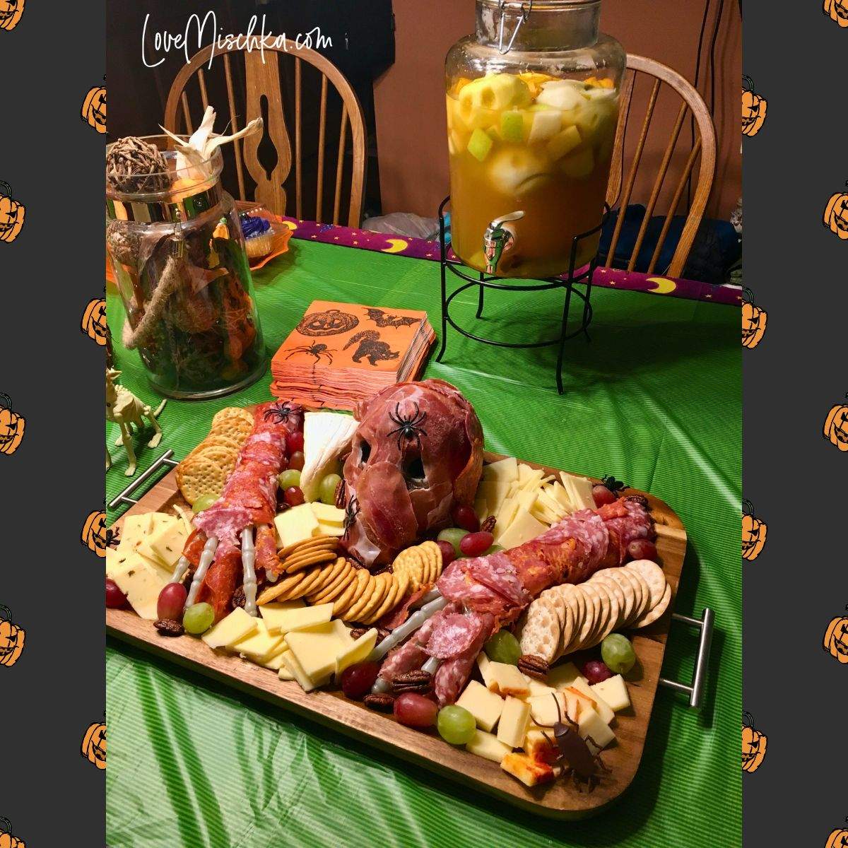 A skeleton charcuterie board next to a pitcher of apple cider with shrunken apple heads in it on a table with a festive green Halloween table cloth.