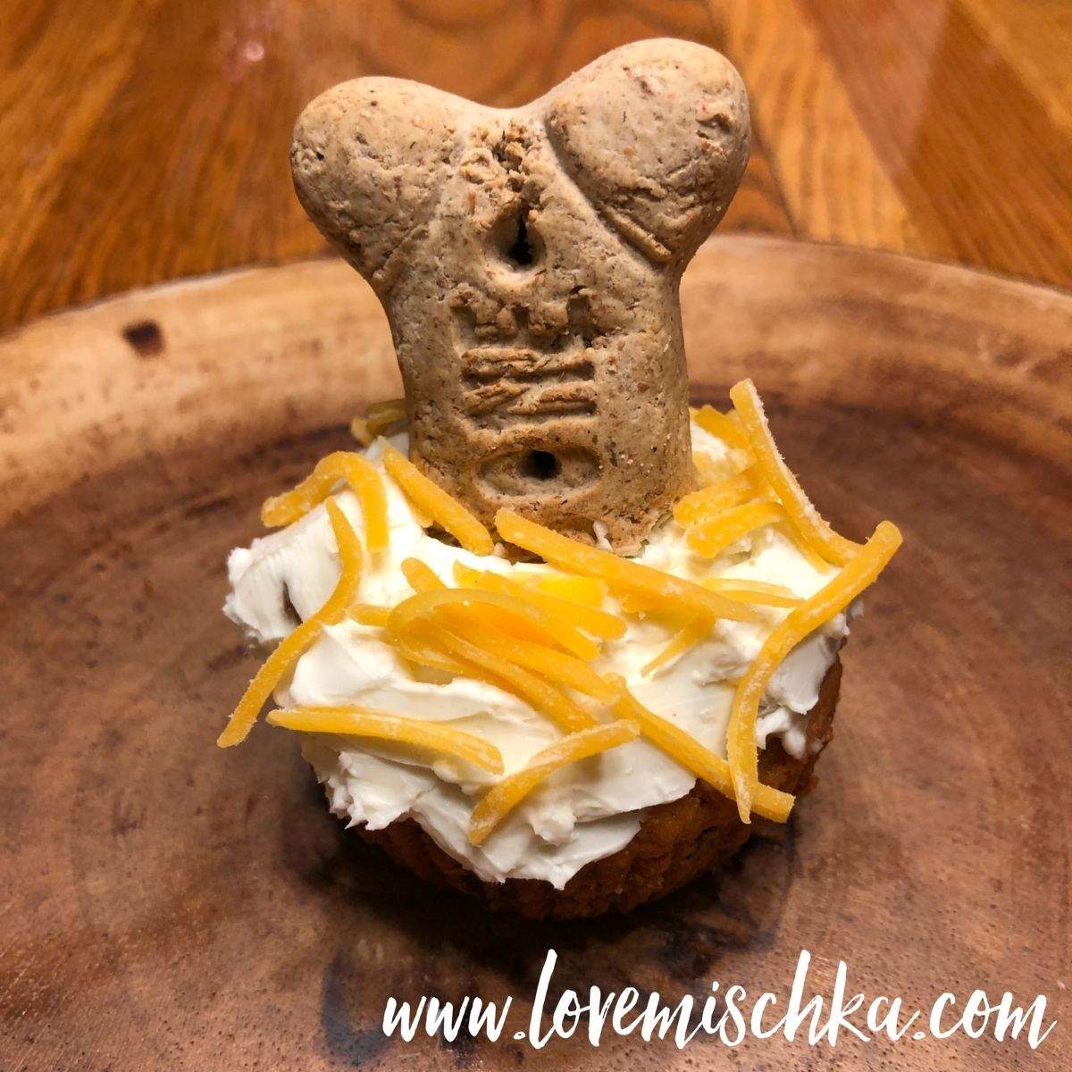 A Dog Cupcake topped with white cream cheese, yellow shredded cheddar cheese, and a big, tan milkbone treat.