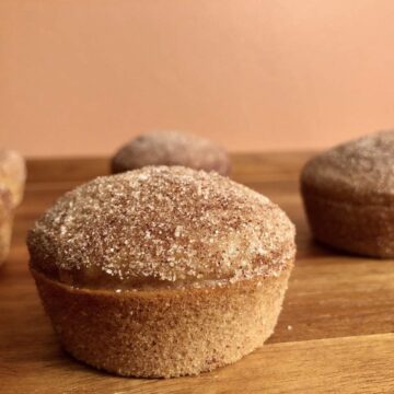 A soft, fluffy Apple Cider Doughnut Muffin with Cinnamon and Sugar on top, next to other Apple Cider Muffins on a brown, wooden serving tray.
