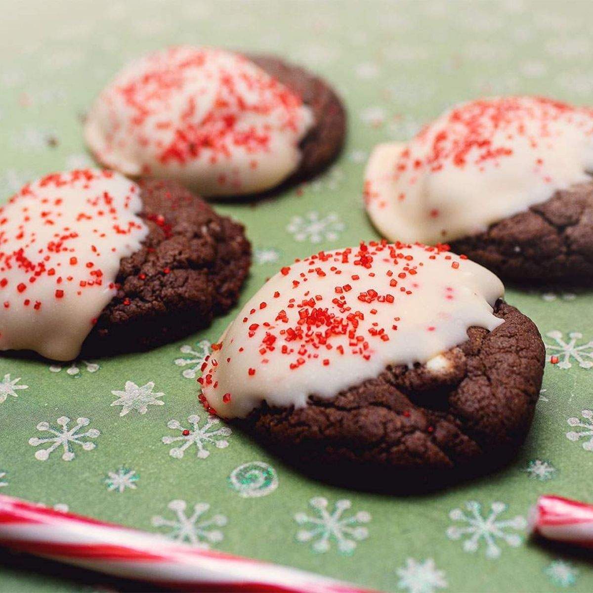 Dark brown chocolate cookies with peppermint kisses, dipped in melted candy cane kisses, sit on a green sheet with snowflakes next to a candy cane.