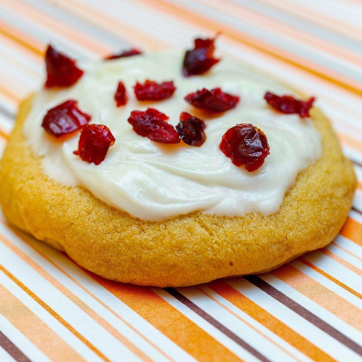 A close up of one round Cranberry Bliss Bar Cookie that is topped with creamy, white cream cheese frosting and pieces of deep red dried cranberries.