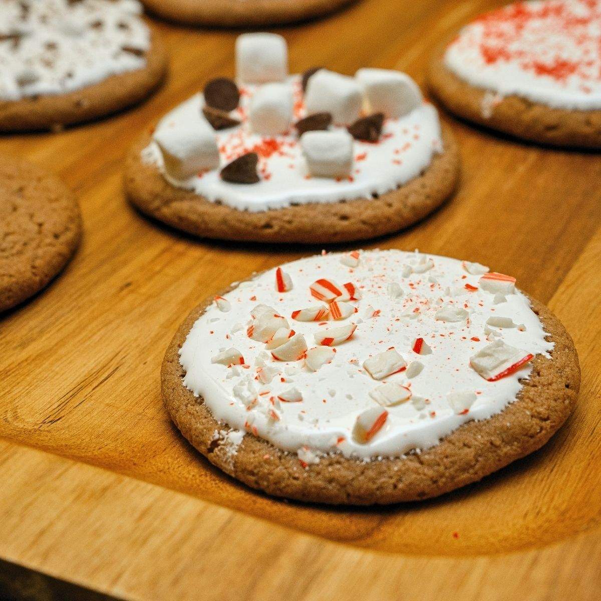 Several brown, round hot chocolate cookies with white marshmallow topping sit on a wooden serving tray. Each cookie has a different, additional topping - crushed candy canes, mini marshmallows, chocolate chips, or holiday sprinkles.