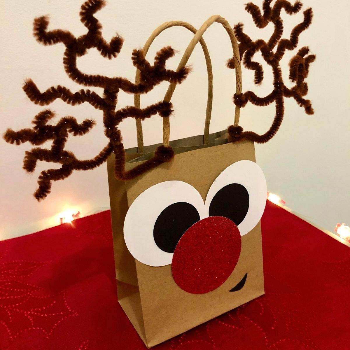 A DIY Reindeer Gift Bag standing up with complex antlers made out of brown pipe cleaners, a large circular nose made out of red glitter cardstock, and big goofy eyes that are cross-eyed.