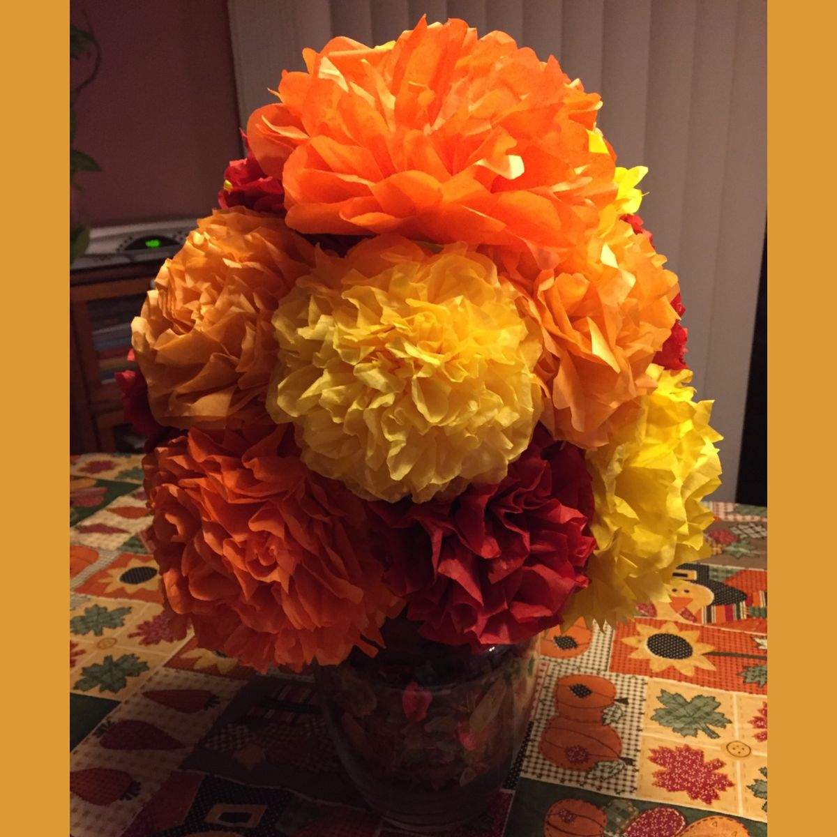 A large bouquet of paper flowers that are made out of different autumn-colored sheets of tissue paper - different shades of orange, yellow, and red. It sits on a kitchen table with a Thanksgiving table cloth.