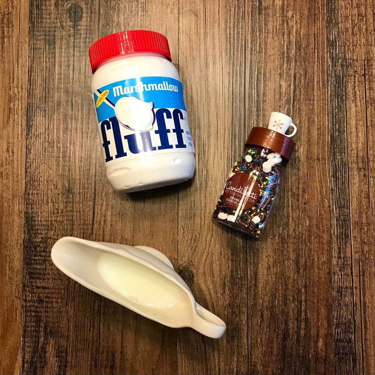 The three ingredients needed to make the fluff marshmallow topping in this hot cocoa cookies recipe - a jar of fluff marshmallow, a small ceramic milk jug of white milk, and a container of fun hot chocolate sprinkles (it even has a 3d hot cocoa mug on top of the lid).