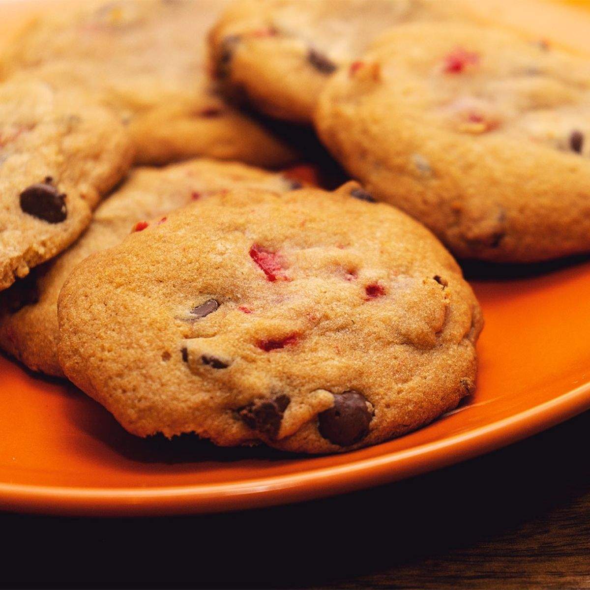 An orange plate of golden chocolate chip cookies with crispy edges, a moist middle, and pieces of bright red maraschino cherries and lots of chocolate chips.