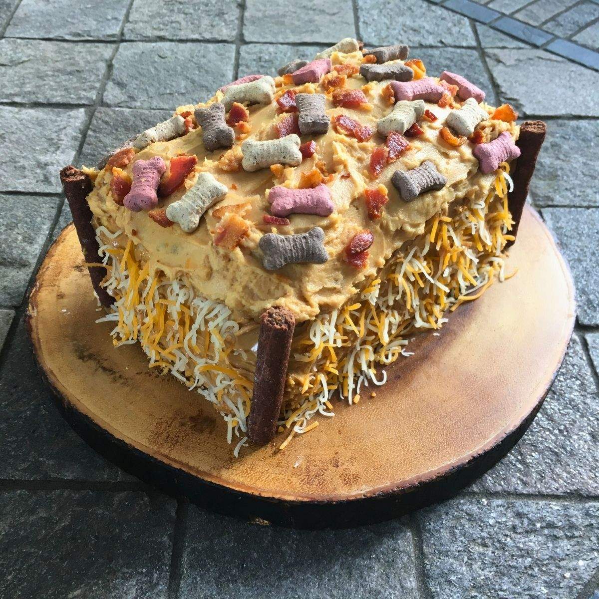 A rectangular cake for dogs with shredded cheese along the sides, dog treats on the corners, and mini bones and bacon pieces on top.
