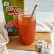 A reddish-orange, iced guava black tea in a glass with a stainless steel straw sits on a tan bamboo cutting board, between a green cartn of guava nectar and two black tea bags.