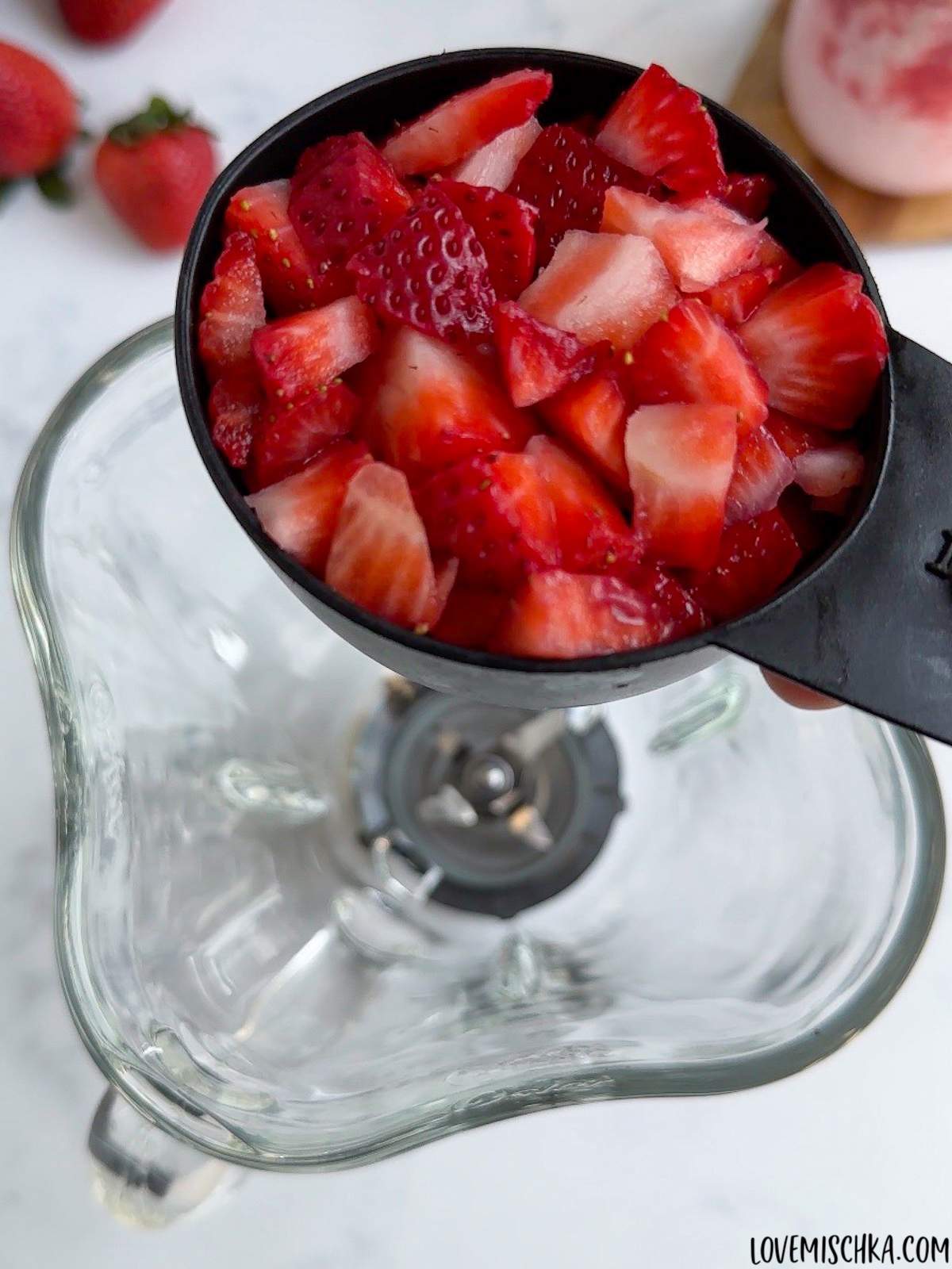 A black measuring cup of bright red, fresh, chopped strawberries is held above a blender in this simple Strawberry Frappuccino Recipe.