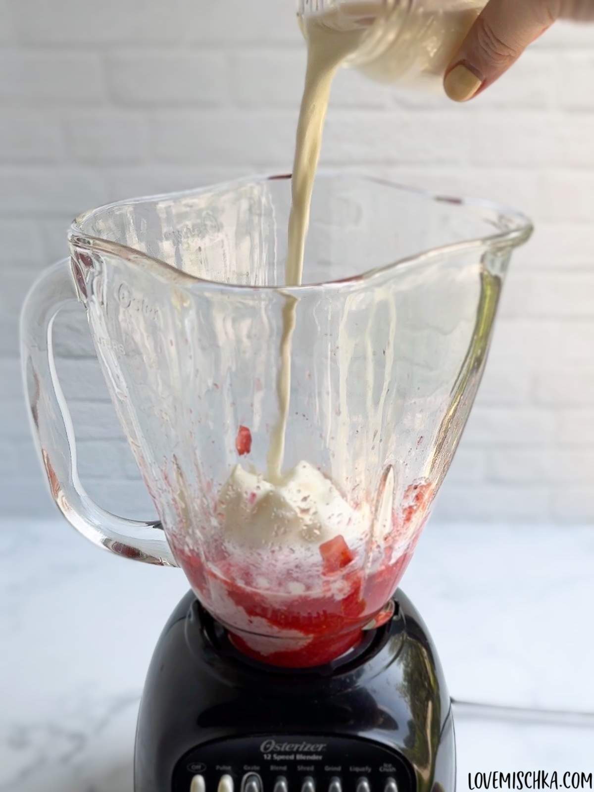 Creamy white cream is poured into a blender that contains homemade strawberry puree and vanilla ice cream in one of the main steps of how to make a strawberry frappuccino.