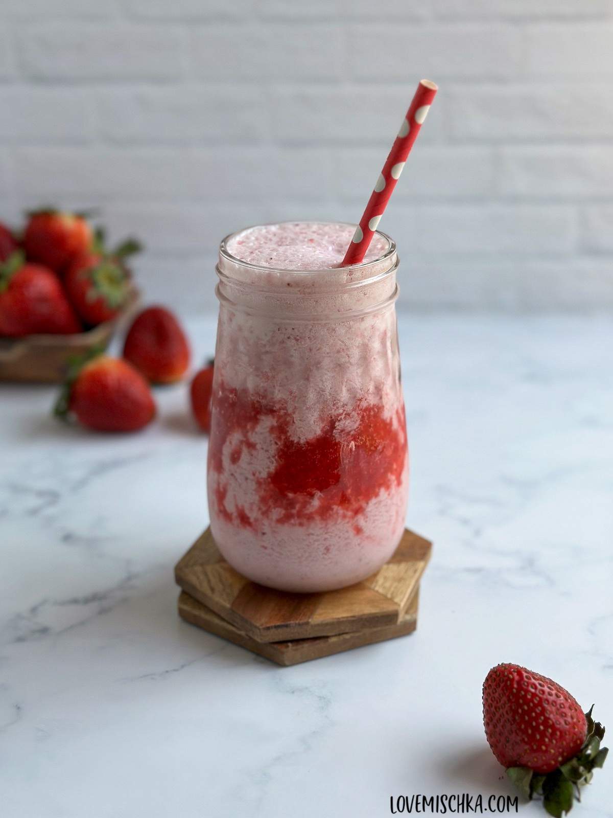 A glass of creamy, pink homemade strawberry frappuccino with fun layers of bright red strawberry puree, sits on two wooden coasters on a marble counter, surrounded by fresh strawberries in this simple drink recipe.