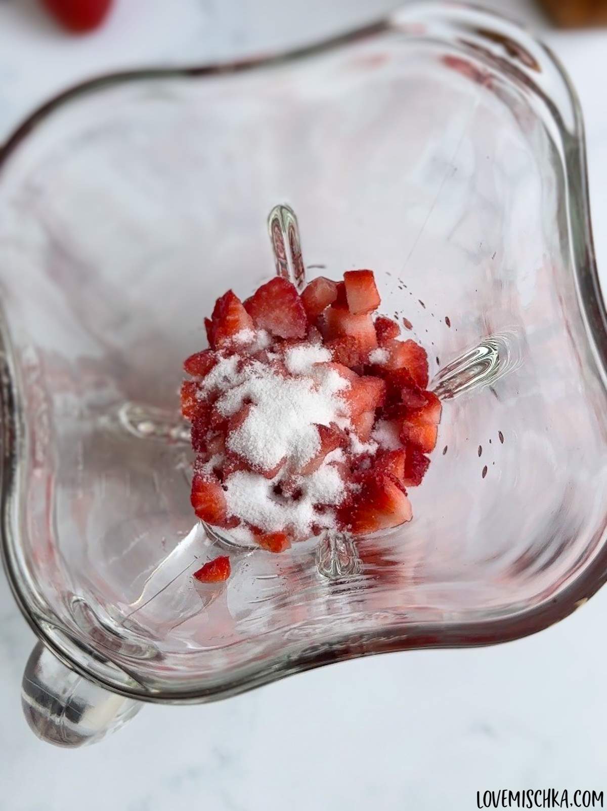 Bright white granulated sugar sits on top of fresh, chopped strawberries in a blender in this homemade strawberry frappuccino recipe.