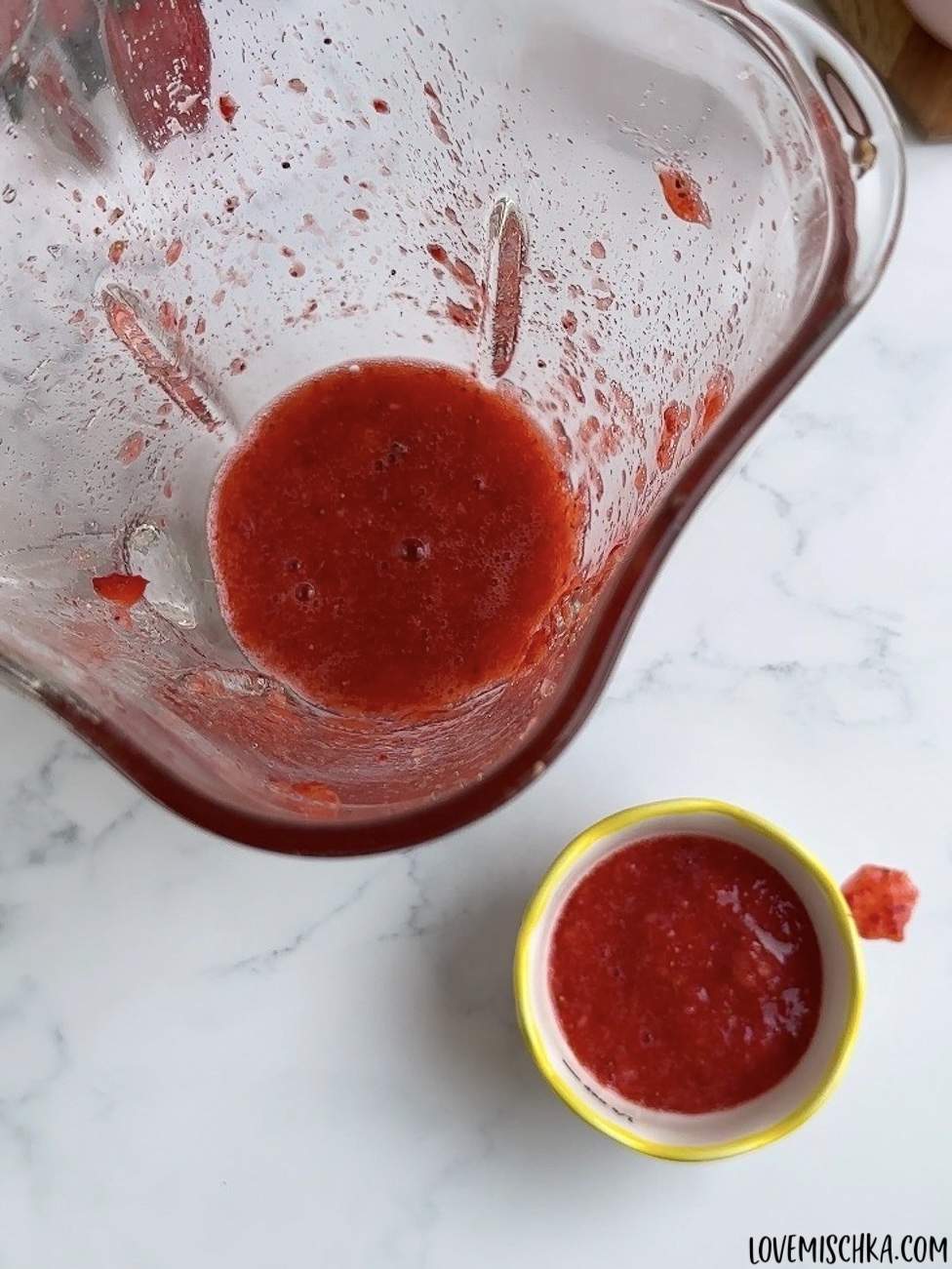 A blender with bright red, homemade strawberry puree that has been blend smooth in this easy Strawberry Frappuccino Recipe and a small bowl of the same strawberry puree being set to the side for later use.