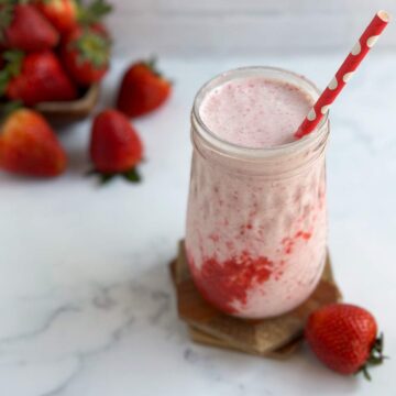 A creamy pink homemade strawberry frappuccino, with a layer of bright red strawberry puree, is in a mason jar glass on two wooden coasters, next to one strawberry in front and lots of fresh strawberries in the back.
