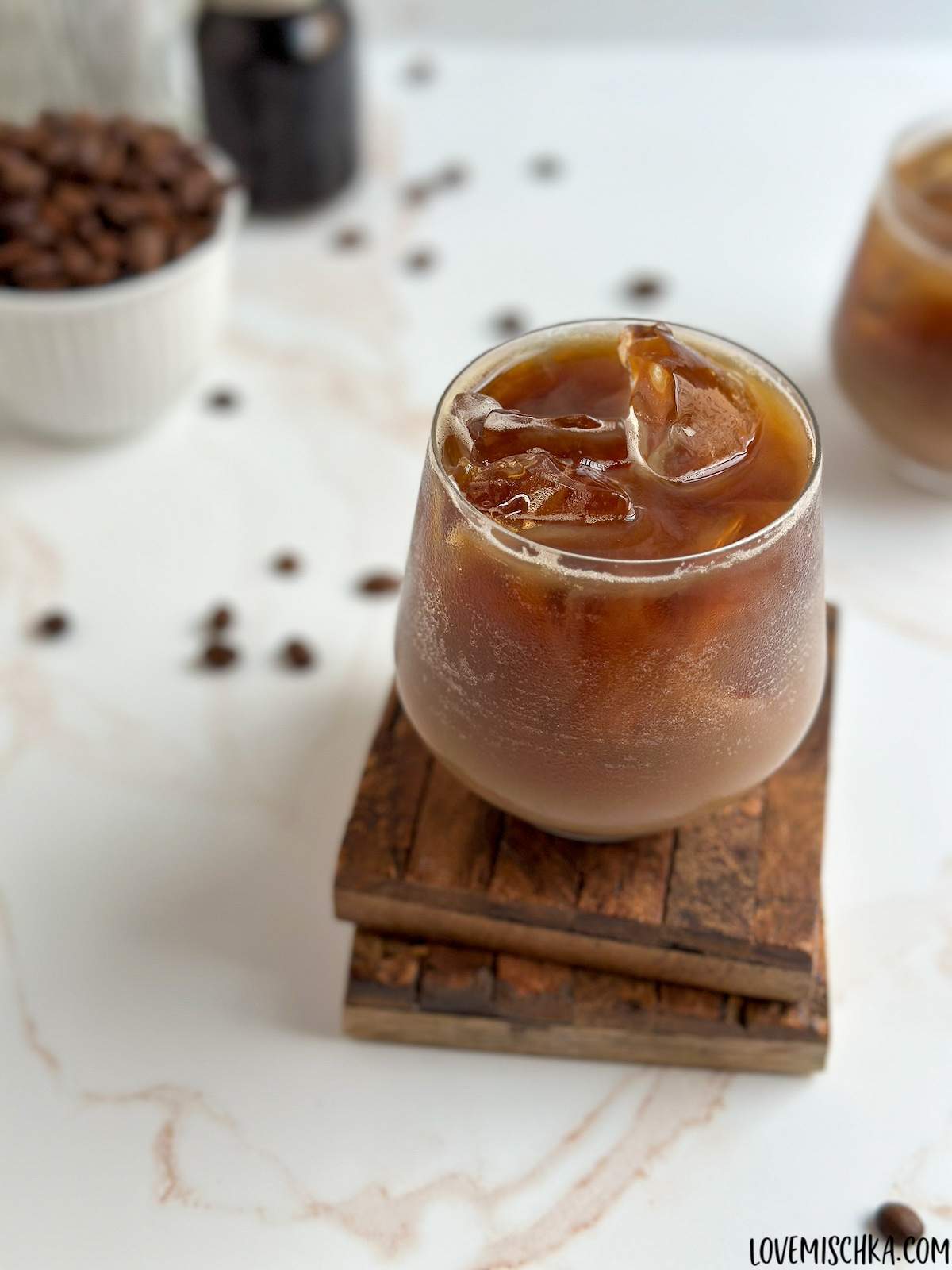 A rich brown Iced Shaken Espresso in a cocktail glass, with refreshing ice cubes, sits on two stacked wooden coasters in front of the simple ingredients in this easy recipe - dark, bold espresso in a small mason jar, whole coffee beans in a small white bowl, and a dairy glass bottle of milk.