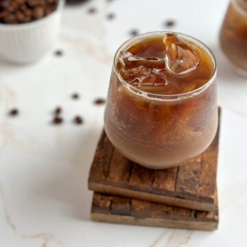 A small glass full of rich brown shaken espresso, with refreshing ice cubes, on two antique wooden coasters and surrounded by loose whole coffee beans in this simple Shaken Espresso Recipe.