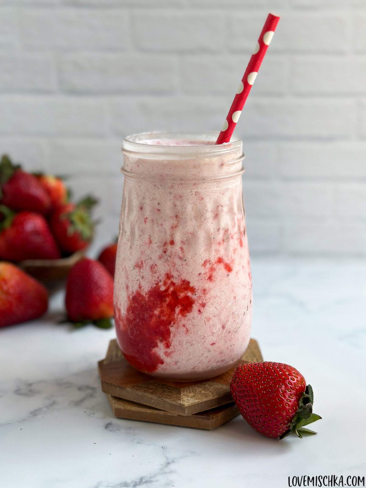 A beautifully pink, creamy Strawberry Frappuccino, with a layer of homemade strawberry puree, sits in front of a bowl of fresh, bright red strawberries in a decorative glass with a red straw with white polka dots.