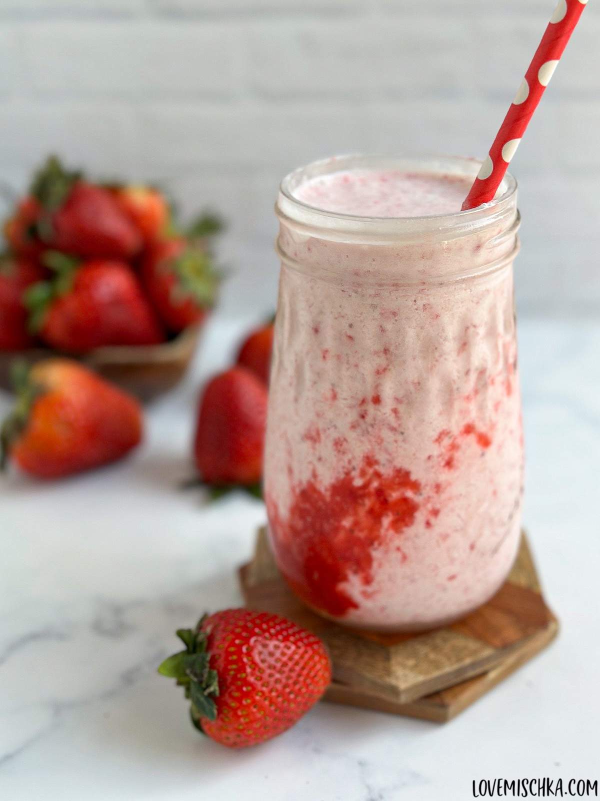 A light pink, creamy, DIY strawberry frappuccino with a layer of bright red homemade strawberry puree, sits in a decorative glass with a red and while polka dot straw. Bright red, fresh strawberries are scattered around the drink in this easy recipe.