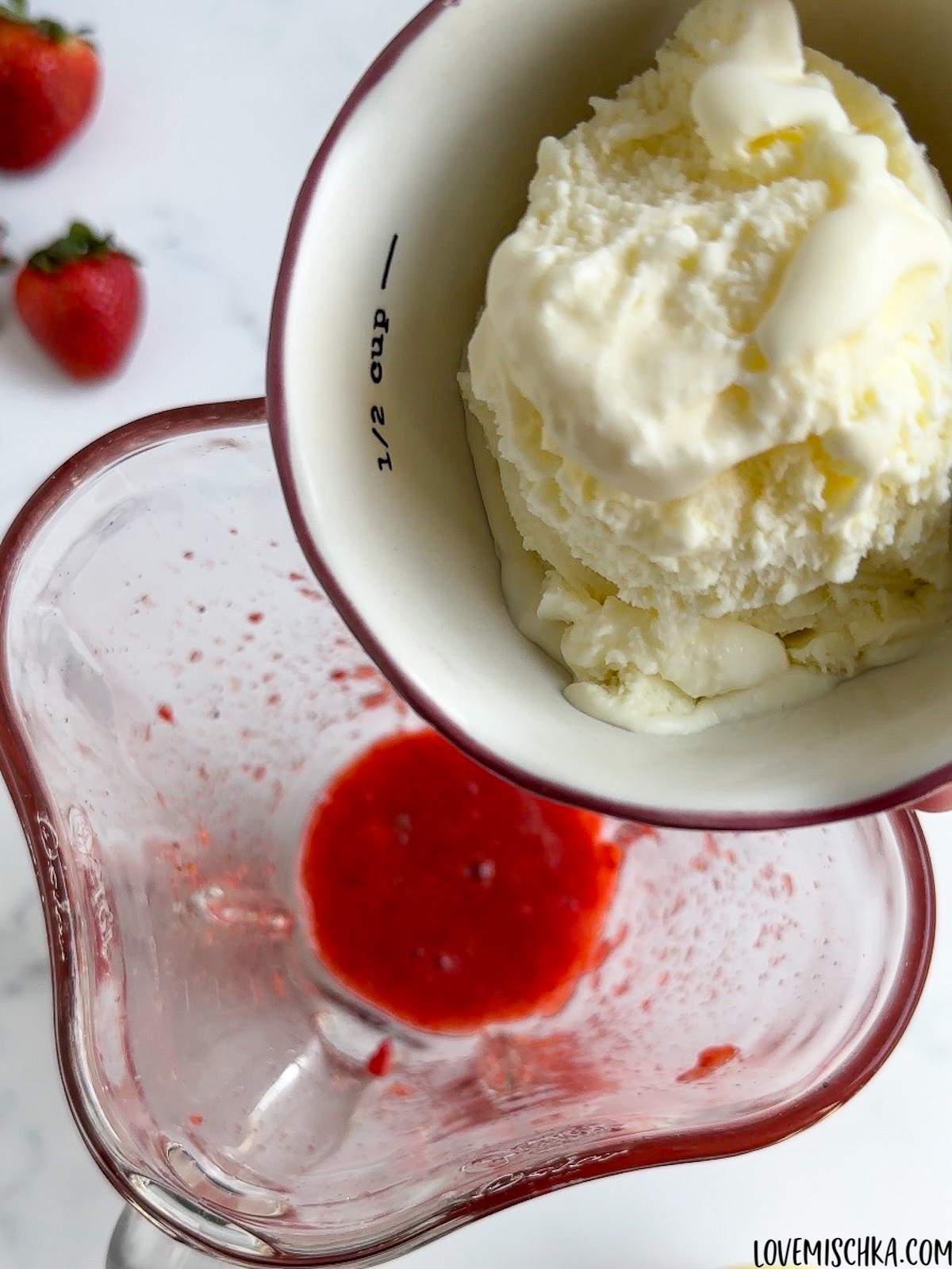 A small bowl of creamy vanilla ice cream is held above bright red, smooth, homemade strawberry puree in a blender in this easy Strawberry Frappuccino Starbucks Recipe.