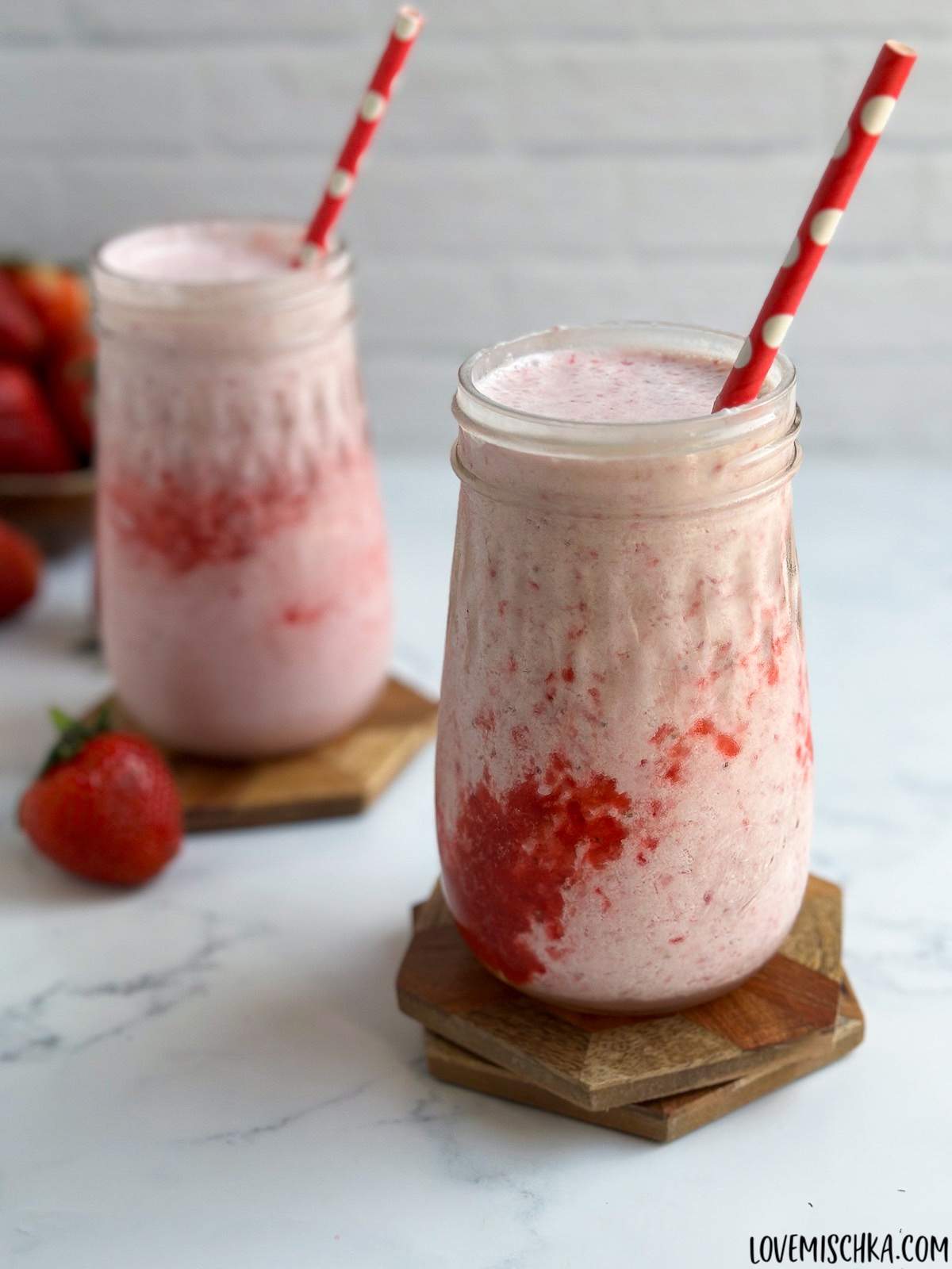 Two beautifully pink, refreshing diy strawberry frappuccinos, in mason jar glasses, sit on wood coasters, with bright red strawberries nearby.