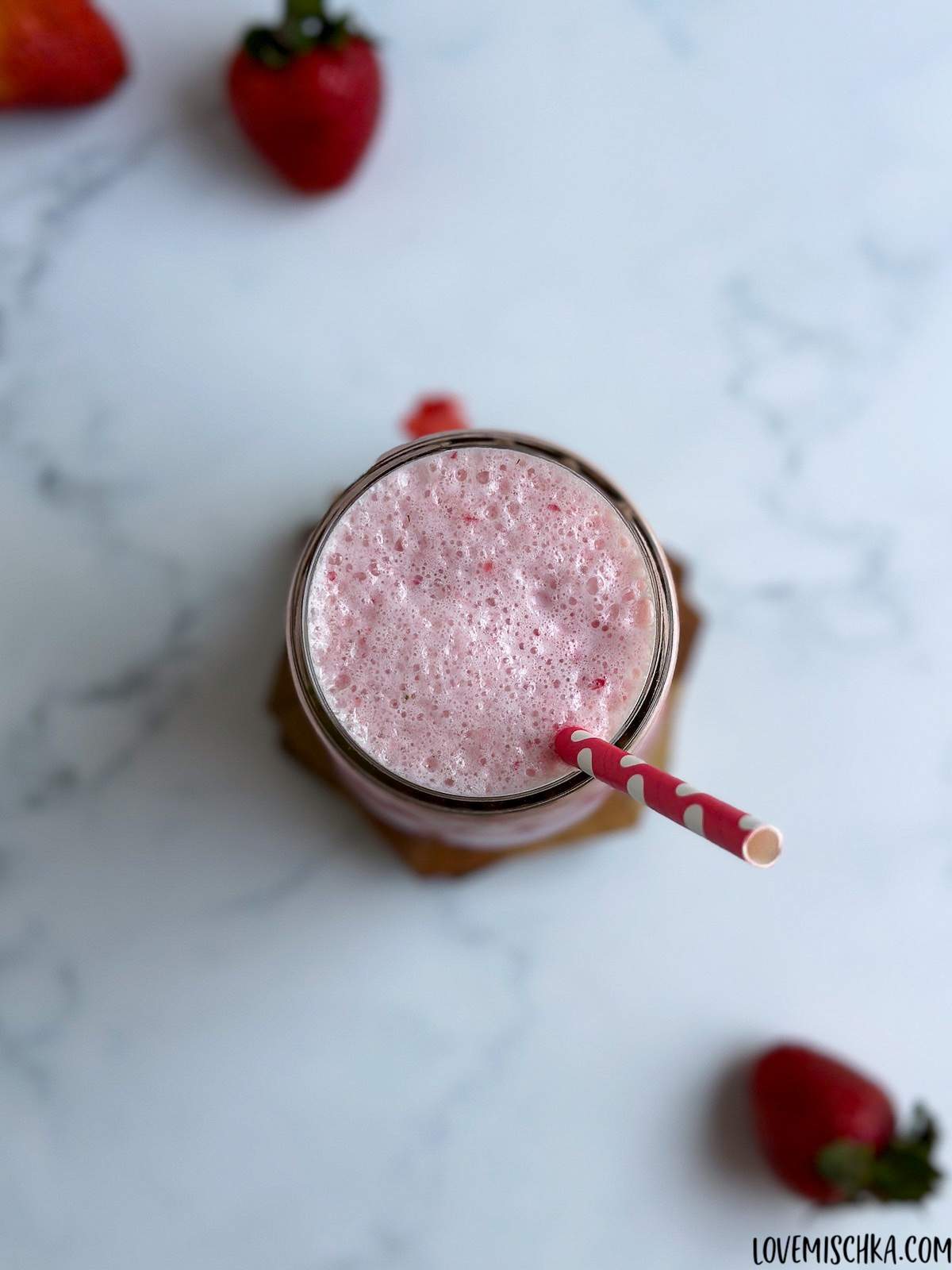 Looking down at a pink, creamy, frothy strawberry frappuccino in a glass with a red paper straw, next to bright red, fresh strawberries.