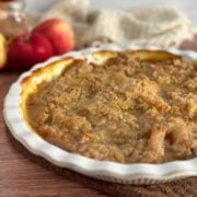 A close up of an Apple Better Pie with crisp, buttery, light golden brown topping in a beautiful white pie plate on a wooden table with bright red apples to the side of it and a beige cloth in the back.