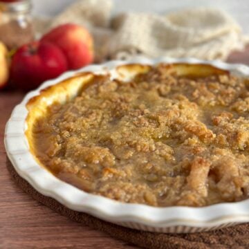 A close up of an Apple Better Pie with crisp, buttery, light golden brown topping in a beautiful white pie plate on a wooden table with bright red apples to the side of it and a beige cloth in the back.